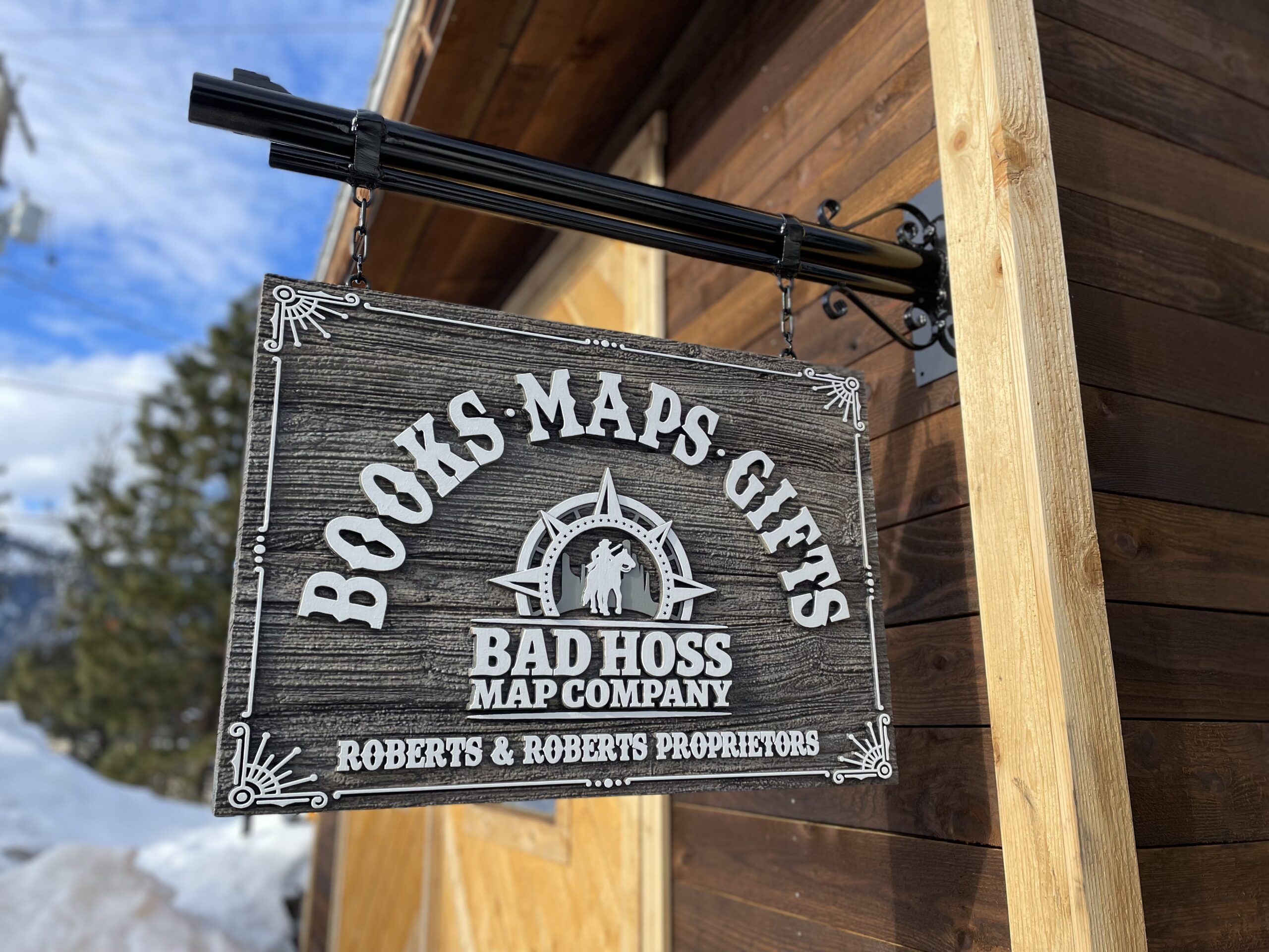 A rustic sign that looks like wood hanging from a metal bracket that looks like a gun barrel.